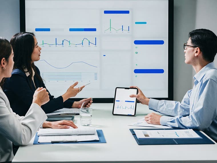 Three managers around a boardroom table facing a wall display of trend charts, one holding a tablet showing the same figures.