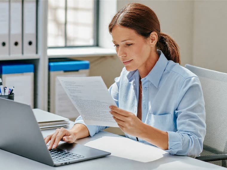 A records officer at her desk reading a printed document against what is open on her laptop, archive boxes on the shelves behind her.